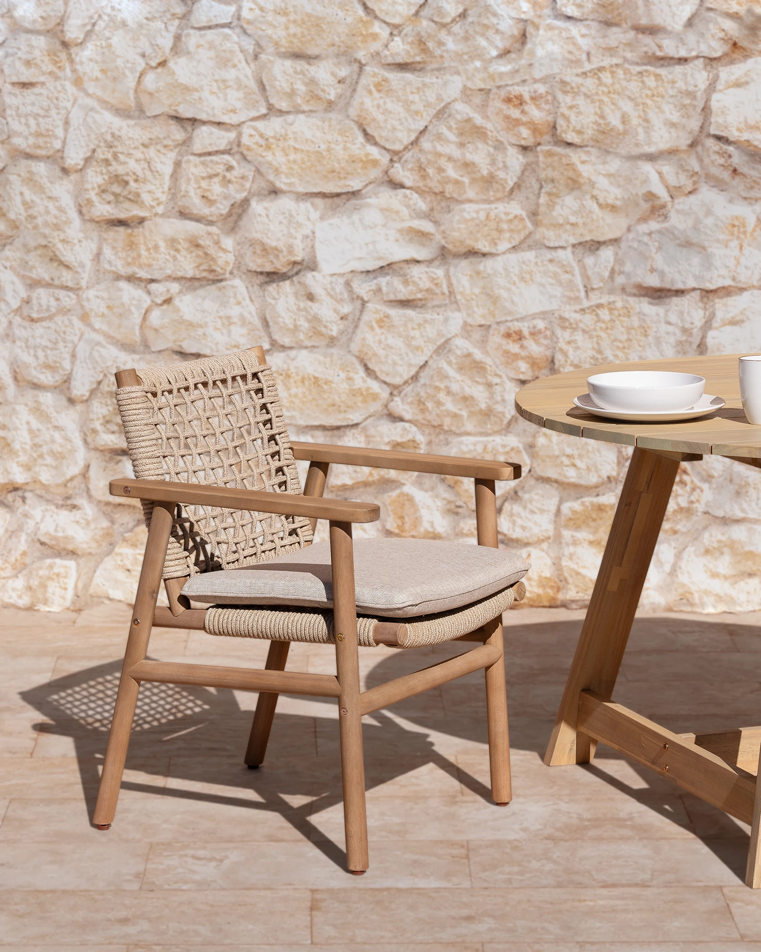 Silla de jardín de madera y cuerda ancha beige con cojín desenfundable color piedra expuesta en exterior con pared de piedra